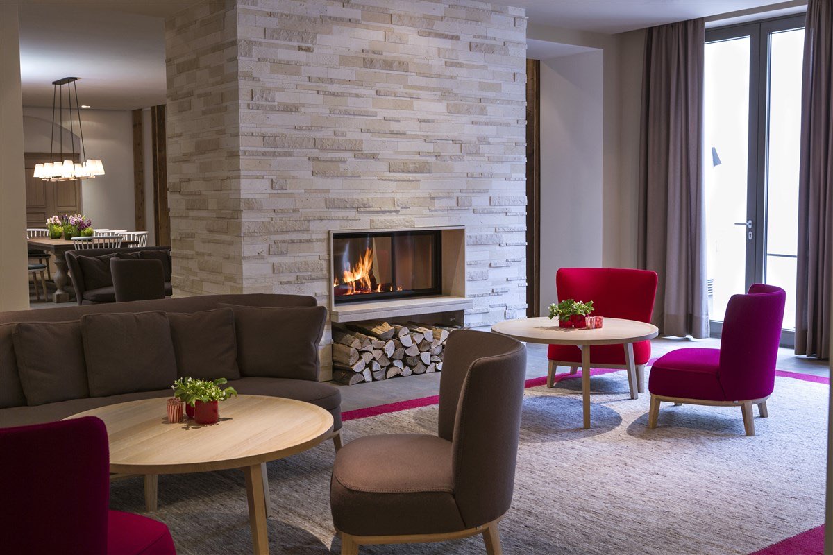 Hotel-/Loungebereich mit in Naturstein verkleidetem Kaminpfeiler, Logenische darunter und runden Holztischen; rote und taupefarbene Sessel