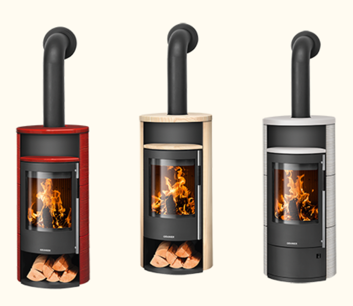 Zylindrischer Ofen mit sandfarbenem, steinartigem Mantel und schwarzer, gebogener Sichtscheibe. Oberhalb der Feuerraumtür befindet sich eine kleine, offene Nische; das Design wirkt warm und wohnlich.