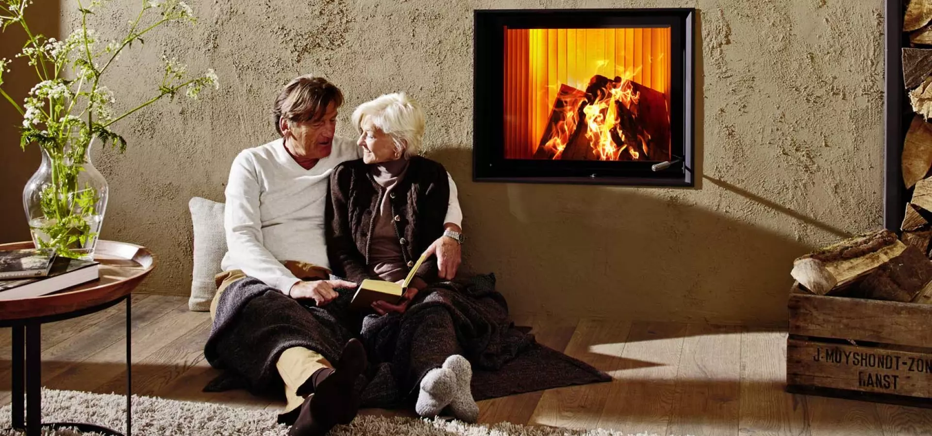 Älteres Paar sitzt auf einer Decke vor einer sandfarbenen Putzwand mit eingelassenem Kaminfeuer; links ein Tisch mit Glasvase, rechts eine Holzkiste mit Brennholz – warme, gemütliche Stimmung